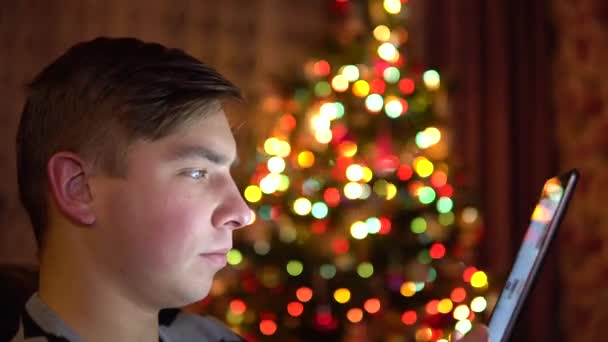 한 젊은이가 크리스마스 트리 뒤에 손에 타블렛을 들고 앉아 있습니다. 방은 크리스마스 분위기로 장식되어 있다. 현대 전자 타블렛. 클로즈업 — 비디오