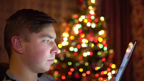 Un joven se sienta con una tableta en las manos sobre el fondo de un árbol de Navidad. La habitación está decorada y llena de ambiente navideño. Tableta electrónica moderna. Primer plano. — Vídeos de Stock