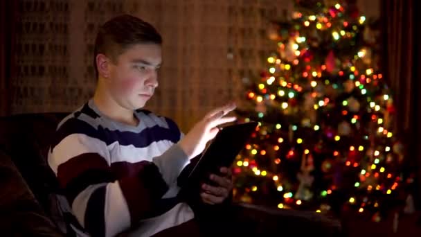 Mladý muž sedí s tabulkou v ruce na pozadí vánočního stromku. Pokoj je zařízený a plný vánoční nálady. Moderní elektronická tableta. — Stock video