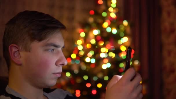 若い男がクリスマスツリーの背景に彼の手にタブレットで座っています。部屋はクリスマスムードで飾られ、記入されています。現代の電子タブレット。閉鎖 — ストック動画