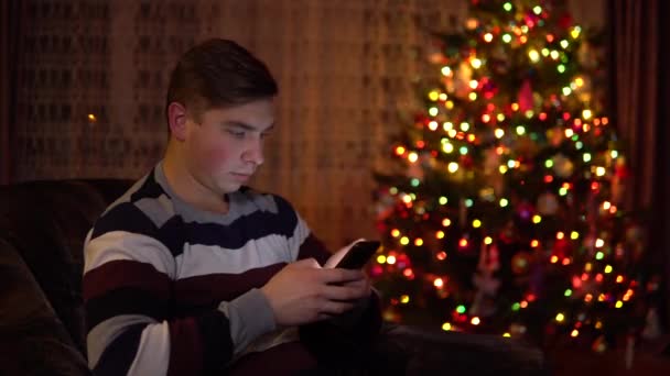 Un joven se sienta con una tableta en las manos sobre el fondo de un árbol de Navidad. La habitación está decorada y llena de ambiente navideño. Tableta electrónica moderna . — Vídeo de stock