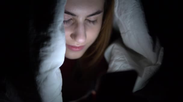 若い女性は携帯電話を手に毛布の下に横たわっている。暗闇の中の女性はパッドを使用します。現代の電子タブレット。閉鎖 — ストック動画