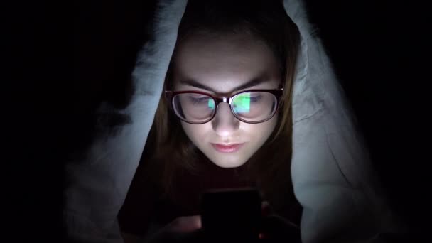 Μια νεαρή γυναίκα ξαπλώνει κάτω από μια κουβέρτα με ένα τηλέφωνο στα χέρια της. Μια γυναίκα στο σκοτάδι χρησιμοποιεί ένα μαξιλάρι. Σύγχρονη ηλεκτρονική ταμπλέτα. Κοντινό πλάνο — Αρχείο Βίντεο