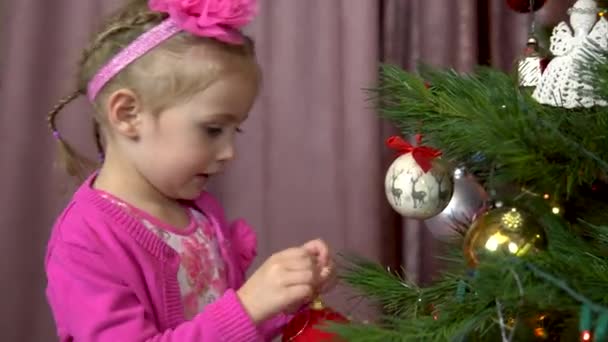 Маленька дівчинка прикрашає ялинку. Дитина вішає м'яч на гілку ялинки. Різдвяна ялинка прикрашена різдвяними іграшками та вогнями . — стокове відео