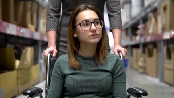 Une jeune femme est assise dans un fauteuil roulant. Une assistante conduit une femme en fauteuil roulant. Marcher le long de la boutique — Video