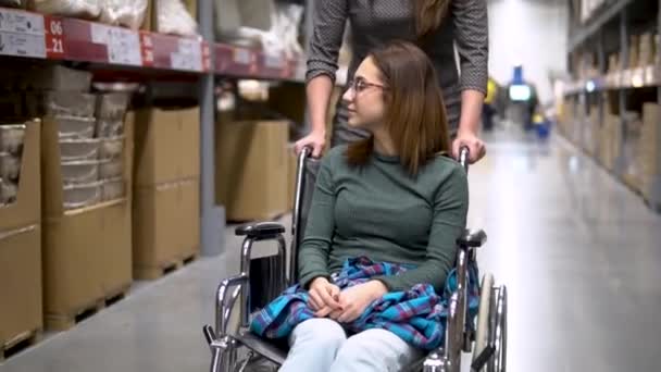 Een jonge vrouw zit in een rolstoel. Vrouwelijke assistente rijdt een vrouw in een rolstoel. Wandelen langs de winkel — Stockvideo