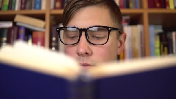 A young man is reading a book in a library. A man with glasses carefully looks at the book closeup. In the background are books on bookshelves. Book library. — 비디오