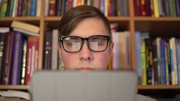 若い男がタブレットで本を読んでいる。眼鏡の男が注意深くタブレットを見ている。背景には本棚の本があります。図書館. — ストック動画