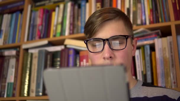 Ein junger Mann liest in einem Tablet ein Buch. Ein Mann mit Brille blickt sorgfältig auf das Tablet. Im Hintergrund stehen Bücher im Bücherregal. Bücherbibliothek. — Stockvideo