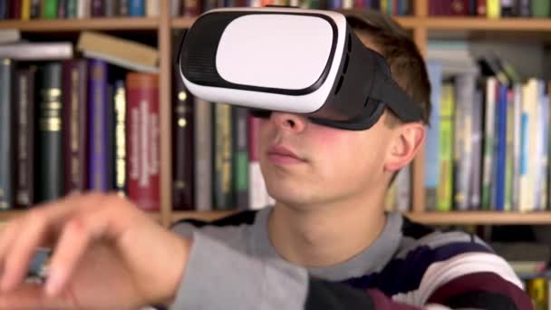 Kütüphanede VR gözlüklü genç adam. Kafasında video kaskı olan bir adam sanal gerçekliği inceler ve ona dokunur. Arka planda kitap raflarında kitaplar var. Kitap kitaplığı. — Stok video
