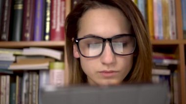 Genç bir kadın tablette bir kitap okuyor. Gözlüklü bir kadın tablete dikkatlice bakar. Arka planda kitap raflarında kitaplar var. Kitap kitaplığı.