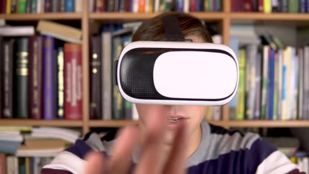 Jeune homme en lunettes VR à la bibliothèque. Un homme avec un casque VR sur la tête examine et touche la réalité virtuelle. En arrière-plan se trouvent des livres sur les étagères. Bibliothèque de livres . — Video