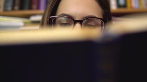 Une jeune femme lit un livre dans une bibliothèque. Une femme avec des lunettes regarde attentivement le livre en gros plan. En arrière-plan se trouvent des livres sur les étagères. Bibliothèque de livres . — Video