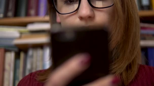 若い女性が図書館でスマートフォンを見ている。メガネの女性は慎重に電話のクローズアップを見ています。背景には本棚の本があります。図書館. — ストック動画