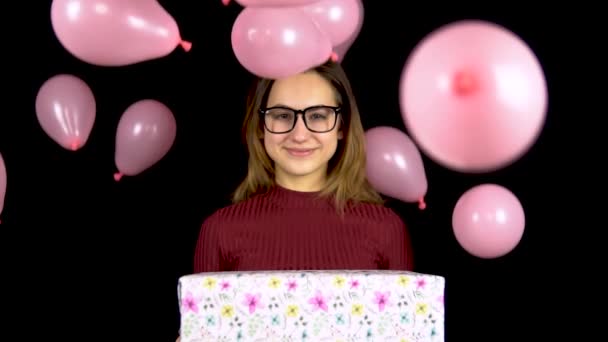 A young girl is standing with a gift, and balloons are falling on her. Smiling woman holding a gift in her hands on a black background. Valentines Day is the day of lovers. Slow motion. — Stock Video