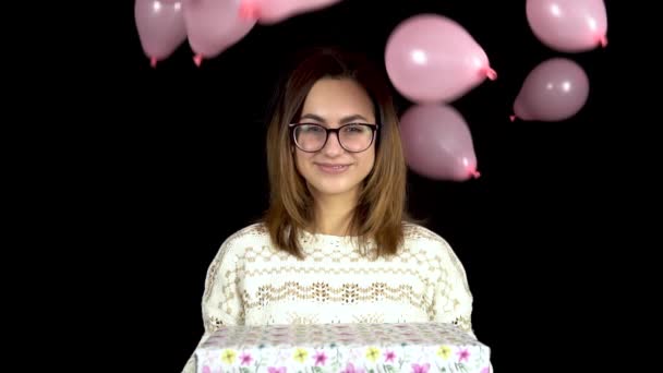 A young woman is standing with a gift, and balloons are falling on her. Smiling woman holding a gift in her hands on a black background. Girl in holiday clothes. Slow motion — Stock Video
