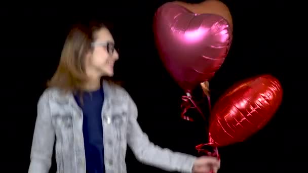 Una joven está de pie con globos en forma de corazón. Mujer saltando de alegría con bolas con helio en las manos sobre un fondo negro. Día de San Valentín es el día de los amantes . — Vídeos de Stock