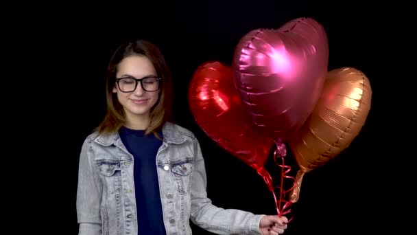 Una joven está de pie con globos en forma de corazón. Mujer sostiene bolas de helio en sus manos sobre un fondo negro. Día de San Valentín es el día de los amantes . — Vídeos de Stock