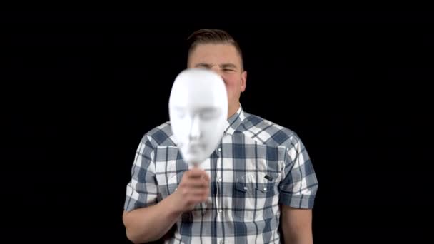 A young man takes off his mask and shows an emotion of disgust on his face. The man is unpleasant. A man hides his face behind a white mask on a black background — Stock Video