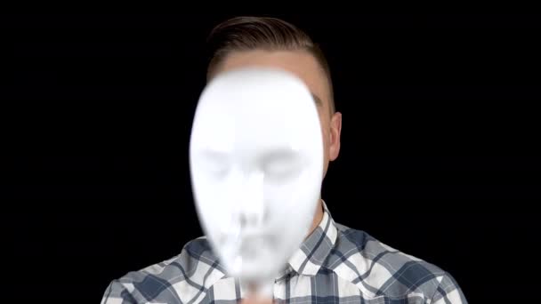 Nimmt ein junger Mann seine Maske ab und zeigt ein Gefühl der Freude auf seinem Gesicht. lächelt der Mann. ein Mann versteckt sein Gesicht hinter einer weißen Maske auf schwarzem Hintergrund — Stockvideo