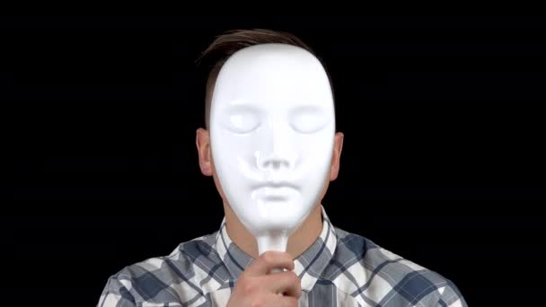 Ein junger Mann nimmt seine Maske ab und zeigt ein Gefühl der Traurigkeit im Gesicht. Trauriger Mann im Hemd. ein Mann versteckt sein Gesicht hinter einer weißen Maske auf schwarzem Hintergrund. — Stockvideo