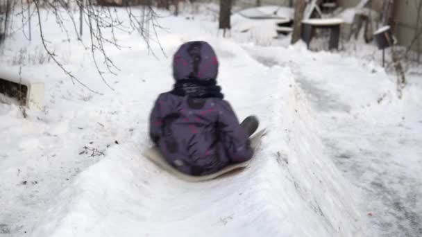 A child rolls on a snow slide. Winter fun on the street. In the cold winter, the child is dressed in a jacket with a scarf. Back view — Stock Video