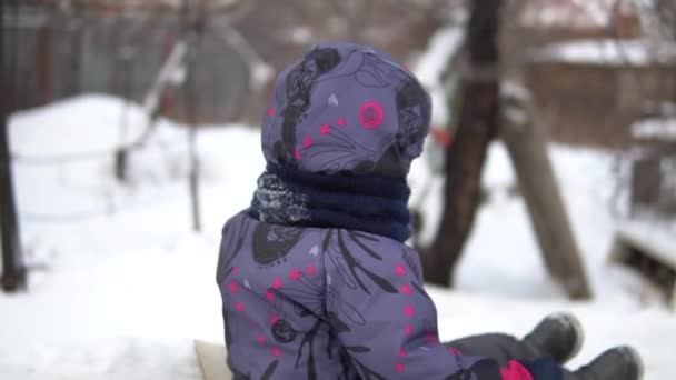 Uma criança rola em um deslizamento de neve. Diversão de inverno na rua. No inverno frio, a criança está vestida com um casaco com um cachecol. Vista lateral. Movimento lento — Vídeo de Stock