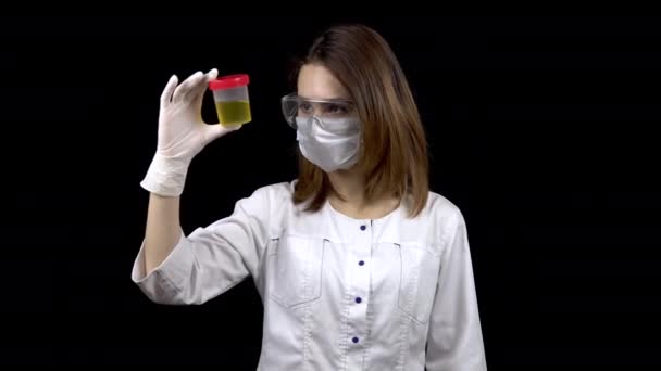 Una joven doctora está revisando las pruebas de orina. Una mujer examina un frasco de prueba que contiene orina sobre un fondo negro . — Vídeos de Stock