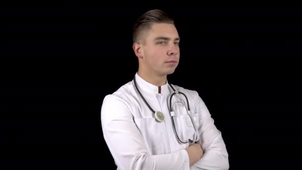 A young male doctor turns his head looking at the camera and smiling. A man stands with a stethoscope on his neck on a black background. — Stock Video