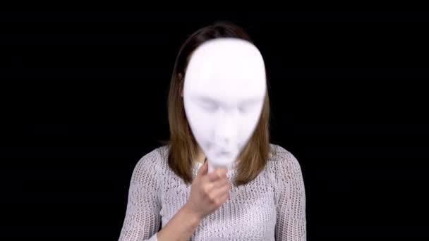 Egy fiatal nő leveszi a maszkot és szomorúságot mutat az arcán. A lány szomorúan néz a kamerába. A nő elrejti arcát egy fehér maszk mögé, fekete alapon.. — Stock videók