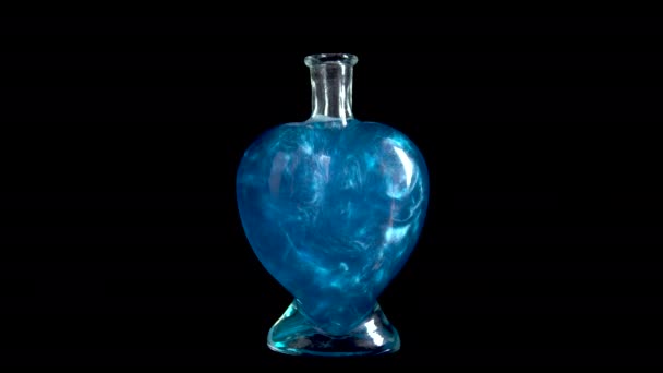 Bottiglia di vetro a forma di cuore con liquido blu. L'elisir sta girando e traboccando di liquido. Pozione d'amore è isolato su uno sfondo nero. — Video Stock