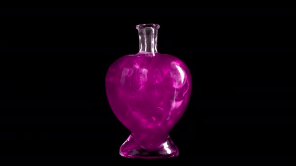 Glass bottle in the form of a heart with pink liquid. The elixir is spinning and overflowing with liquid. Potion of love is isolated on a black background. — Stock Video