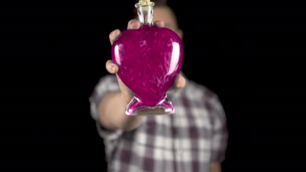 Un joven sostiene una botella con una poción de amor en la cámara. Botella de vidrio en forma de corazón con líquido rosa. Día de San Valentín es el día de todos los amantes . — Vídeo de stock