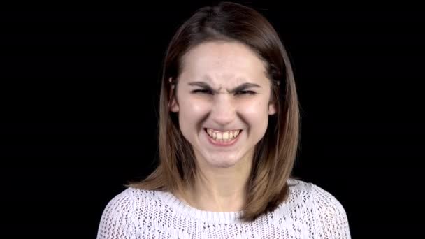 La joven mujer muestra emociones de ira en su cara. Chica sonríe dientes y frunce el ceño cejas sobre un fondo negro primer plano . — Vídeos de Stock