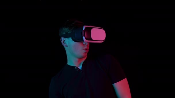 Молода людина в окулярах VR. Чоловік, занурений у віртуальну реальність, робить рухи руками. Блакитне і червоне світло падає на чоловіка на чорному тлі . — стокове відео