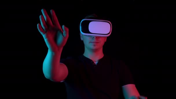 Jeune homme en lunettes VR. Un homme plongé dans la réalité virtuelle fait des mouvements avec ses mains. La lumière bleue et rouge tombe sur un homme sur un fond noir . — Video