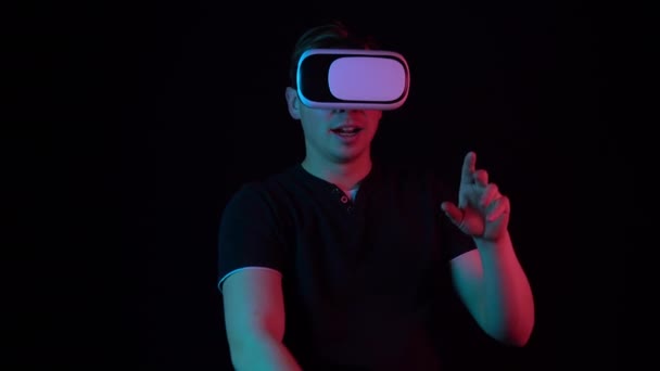 Young man in VR glasses. A man immersed in virtual reality makes movements with his hands. Blue and red light falls on a man on a black background. — Stok video