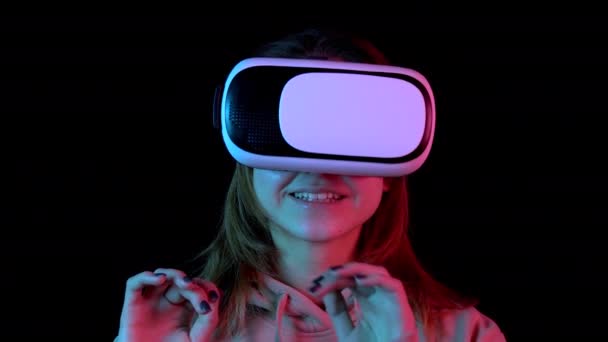 Mujer joven en gafas VR primer plano. Una mujer inmersa en la realidad virtual hace movimientos con las manos. Luz azul y roja cae sobre una mujer sobre un fondo negro . — Vídeo de stock