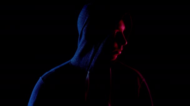 Mladý muž v kapuci se rozhlíží kolem. Útočník stojí ve tmě. Modré a červené světlo dopadá na osobu na černém pozadí. — Stock video