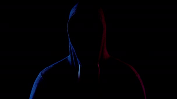 Ένας νεαρός άνδρας στέκεται στο καπό και κρύβει το πρόσωπό του. Ένας εισβολέας στέκεται στο σκοτάδι. Μπλε και κόκκινο φως πέφτει στο πρόσωπο σε μαύρο φόντο. — Αρχείο Βίντεο