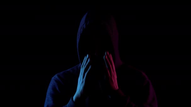 Молодой человек снимает капюшон. Нападавший стоит в темноте. Синий и красный свет падает на человека на черном фоне . — стоковое видео