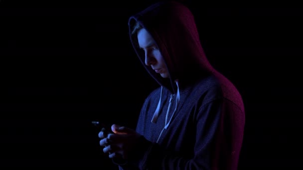 A young man in a hood with a phone in his hands. Hacker makes a hack through the phone. Blue and red light falls on a person on a black background. — 비디오