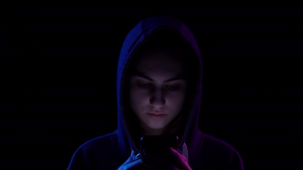 A young woman in a hood with a phone in her hands closeup. Hacker makes a hack through the phone. Blue and red light falls on a person on a black background. — Stok video