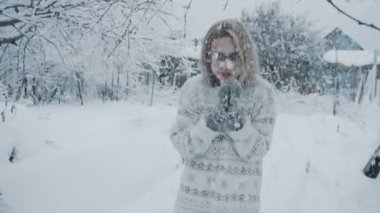 The girl in glasses, in a sweater and mittens stands on the street under the snowfall in winter slow