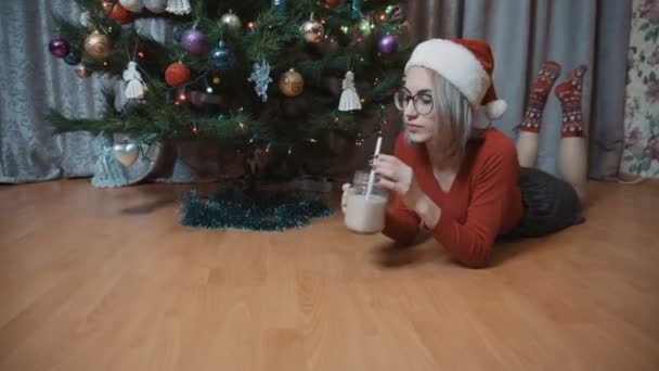 Het meisje ligt op de maag bij de kerstboom en drinkt chocolademelk. Meisje met kerstmuts — Stockvideo
