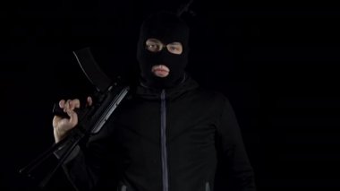 Kar maskeli bir adam AK-47 saldırı tüfeğiyle ayakta duruyor. Haydut omzuna silah dayayıp kameraya nişan alıyor. Siyah bir arkaplanda.