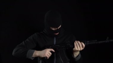 Kar maskeli bir adam AK-47 saldırı tüfeğiyle ayakta duruyor. Haydut makineye saldırır ve durur. Siyah bir arkaplanda.