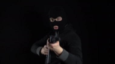 Kar maskeli bir adam AK-47 saldırı tüfeğiyle ayakta duruyor. Haydut otomatik olarak kameraya nişan alır. Siyah bir arkaplanda.