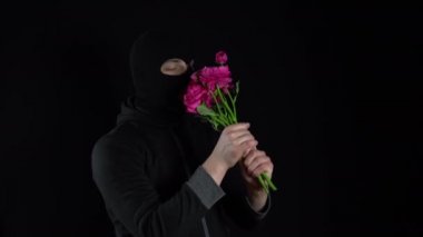 Kar maskeli bir adam elinde çiçeklerle dikiliyor. Haydut, siyah arka planda bir buket pembe çiçeği kokluyor..