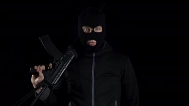 Kar maskeli bir adam AK-47 saldırı tüfeğiyle ayakta duruyor. Siyah bir arka planda, omzunda silahla bir haydut duruyor..
